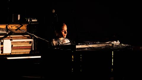 Eine junge Frau sitzt an einem Flügel auf einer Bühne. Der Hintergrund ist überwiegend schwarz, sie sitzt im Licht und wirkt versunken in die Musik. 
