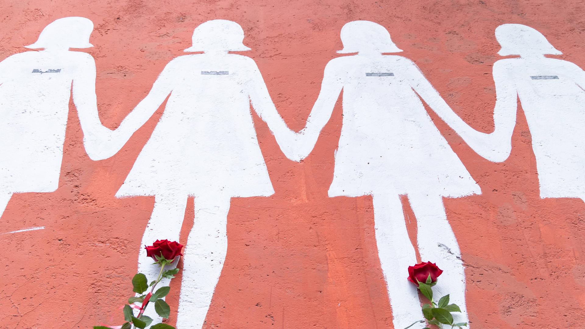 An eine rosa Wand sind weiße Umrisse von Frauen gemalt. Davor wurden rote Rosen abgelegt.
