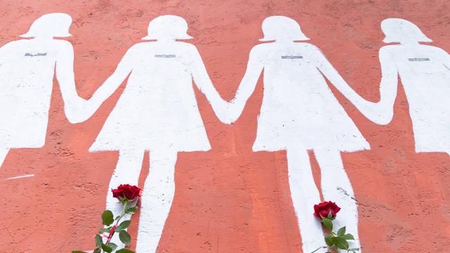 An eine rosa Wand sind weiße Umrisse von Frauen gemalt. Davor wurden rote Rosen abgelegt.