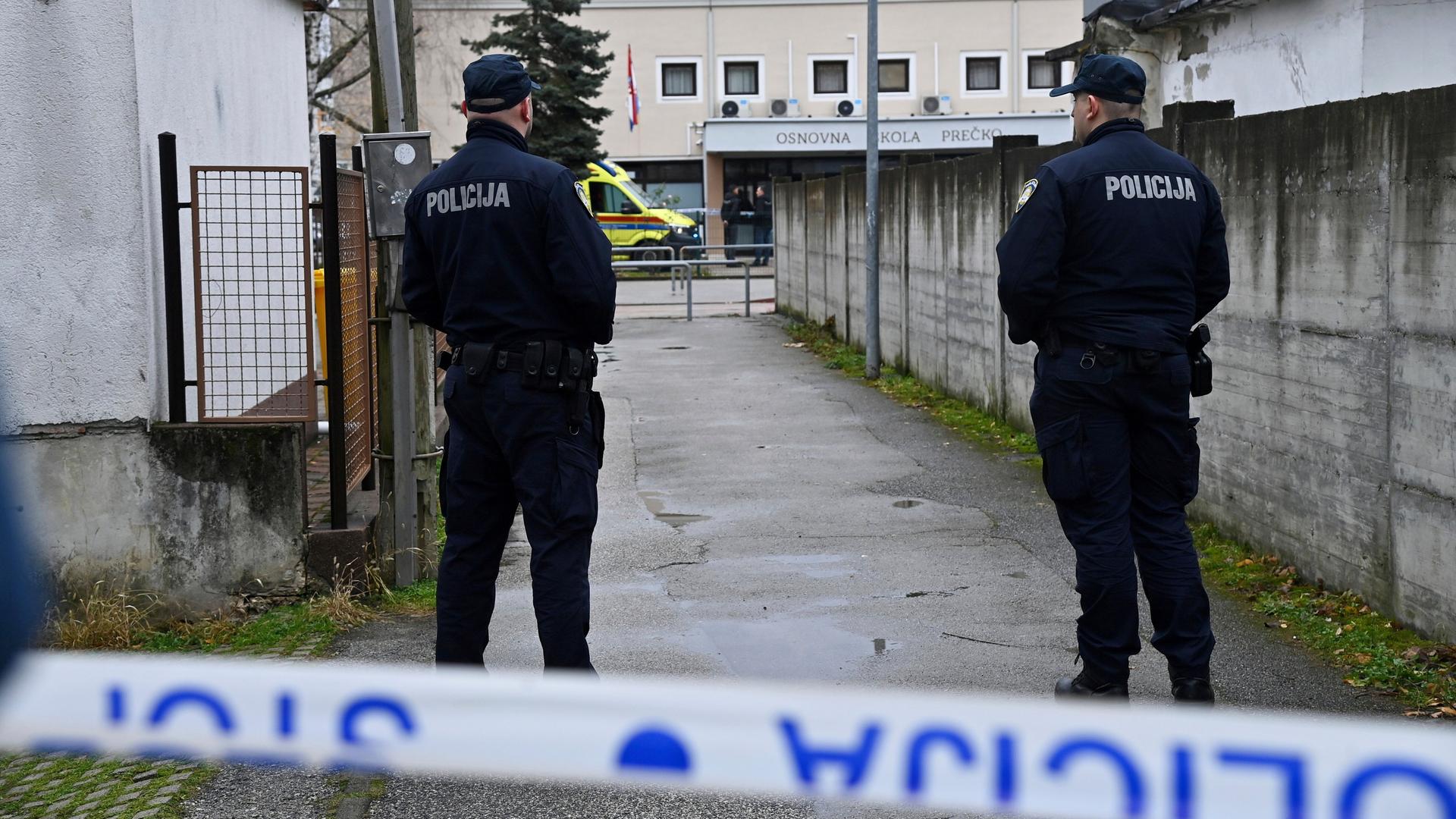 Zwei Polizisten stehen mit dem Rücken zur Kamera. Sie sichern den Zugang zu der Grundschule. Im Hintergrund ist das Schulgebäude zu sehen. Zwei Polizisten stehen mit dem Rücken zur Kamera. Sie sichern den Zugang zu der Grundschule. Im Hintergrund ist das Schulgebäude zu sehen.