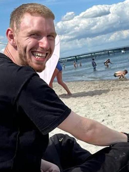 Simon Arnold sitzend am Strand in die Kamera lächelnd 
