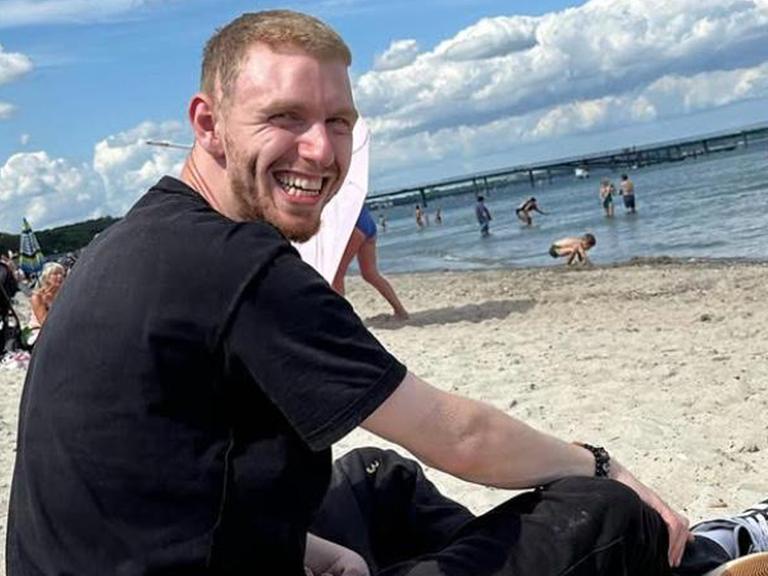 Simon Arnold sitzend am Strand in die Kamera lächelnd 