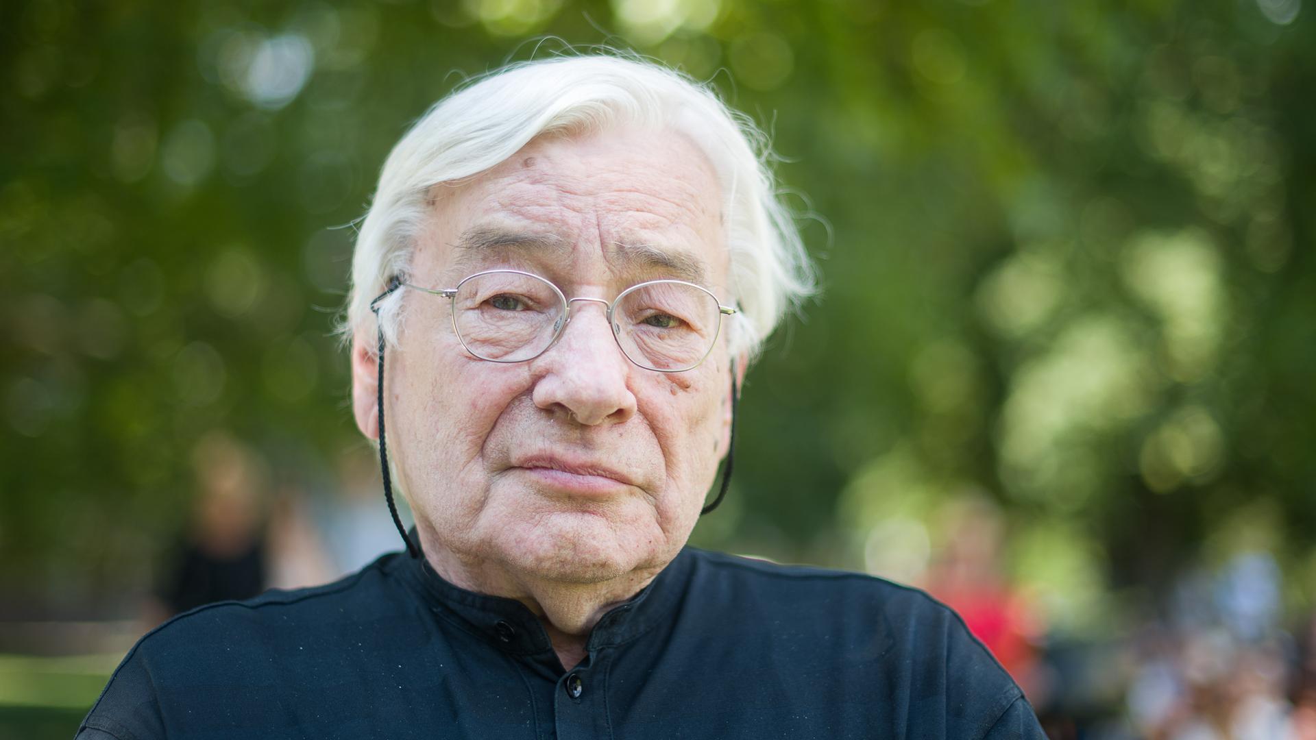 Klaus Zehelein, ehemaliger Intendant der Oper Stuttgart, sitzt mit Brille, weißen Haaren und schwarzem Hemd im Grünen.. Klaus Zehelein, ehemaliger Intendant der Oper Stuttgart, sitzt mit Brille, weißen Haaren und schwarzem Hemd im Grünen..