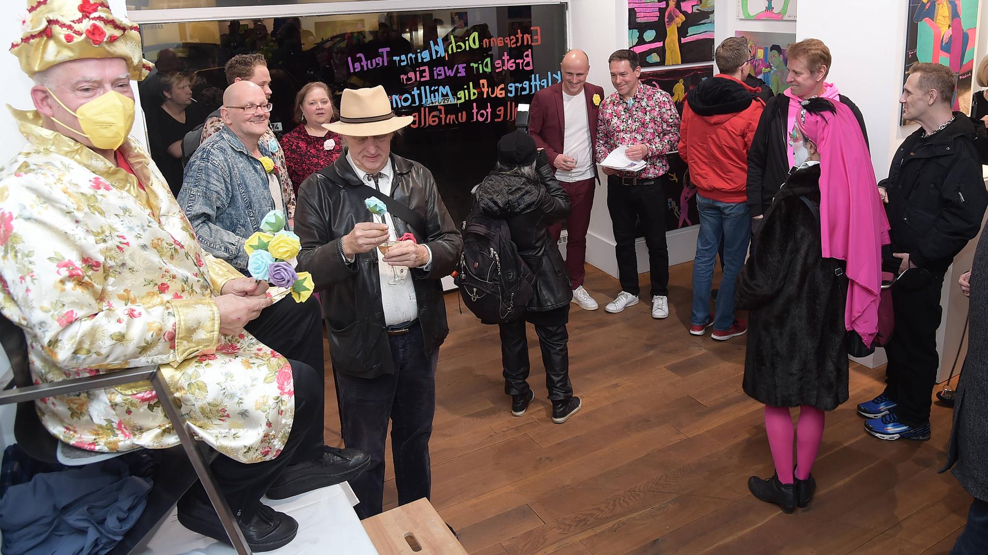 Rosa von Praunheim thront bei der Vernissage seiner Ausstellung in Berlin auf einem Stuhl, der auf einem Tisch steht. Er hat einen Spitzhut auf und ein Gewand in passenden Farben, er trägt eine Maske zum Schutz vor einer Corona-Ansteckung. 