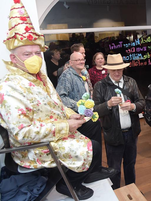 Rosa von Praunheim thront bei der Vernissage seiner Ausstellung in Berlin auf einem Stuhl, der auf einem Tisch steht. Er hat einen Spitzhut auf und ein Gewand in passenden Farben, er trägt eine Maske zum Schutz vor einer Corona-Ansteckung. Rosa von Praunheim thront bei der Vernissage seiner Ausstellung in Berlin auf einem Stuhl, der auf einem Tisch steht. Er hat einen Spitzhut auf und ein Gewand in passenden Farben, er trägt eine Maske zum Schutz vor einer Corona-Ansteckung.