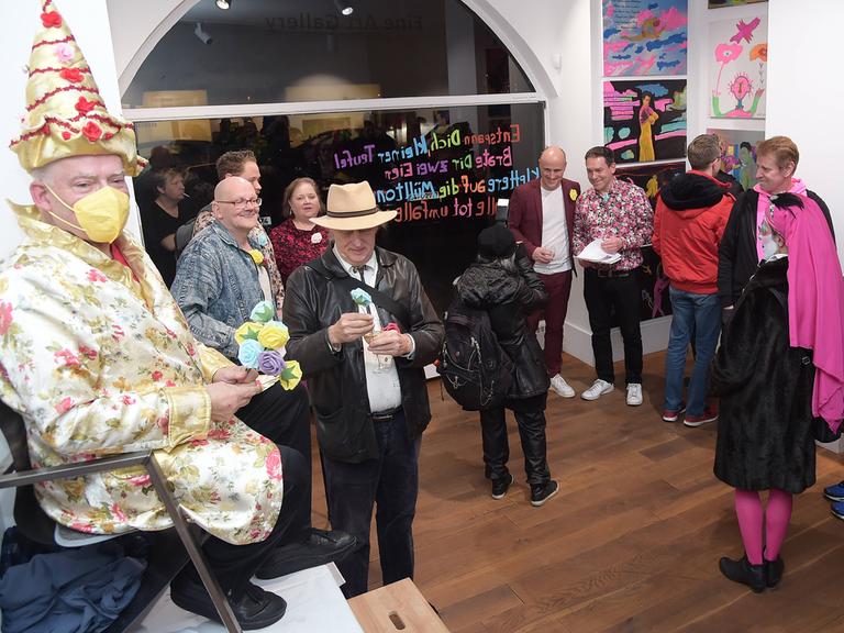 Rosa von Praunheim thront bei der Vernissage seiner Ausstellung in Berlin auf einem Stuhl, der auf einem Tisch steht. Er hat einen Spitzhut auf und ein Gewand in passenden Farben, er trägt eine Maske zum Schutz vor einer Corona-Ansteckung. Rosa von Praunheim thront bei der Vernissage seiner Ausstellung in Berlin auf einem Stuhl, der auf einem Tisch steht. Er hat einen Spitzhut auf und ein Gewand in passenden Farben, er trägt eine Maske zum Schutz vor einer Corona-Ansteckung.