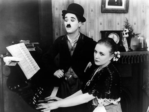 Charlie Chaplin steht in einer Stummfilmaufnahme neben Edna Purviance, die gedankenversunken am Klavier sitzt und spielt. 