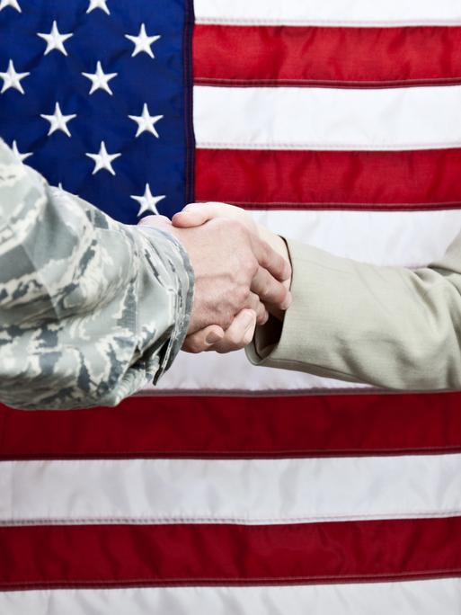 Handschlag zwischen einem Soldaten und einer Zivilistin. Aufnahme vor einer amerikanischen Flagge. Selektiver Fokus auf die Hände.
