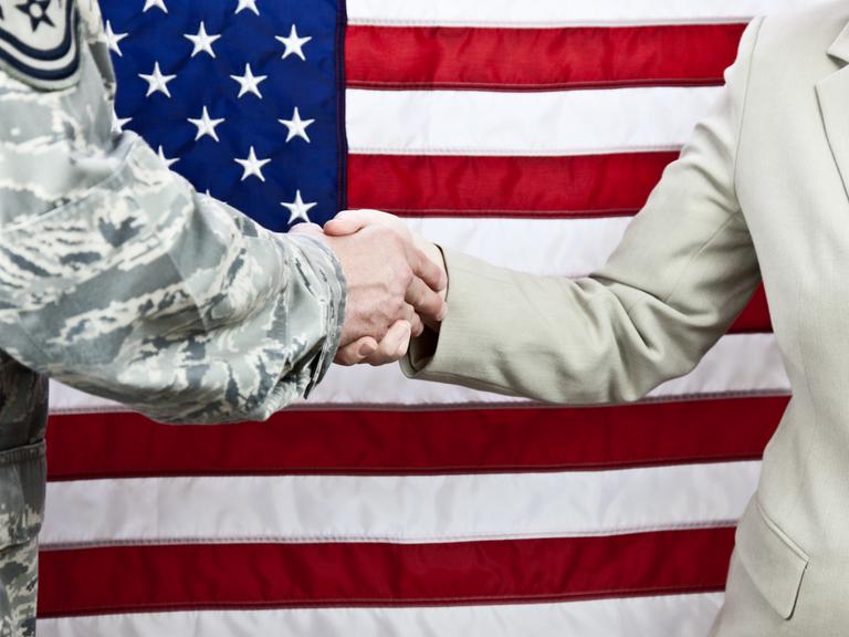 Handschlag zwischen einem Soldaten und einer Zivilistin. Aufnahme vor einer amerikanischen Flagge. Selektiver Fokus auf die Hände.
