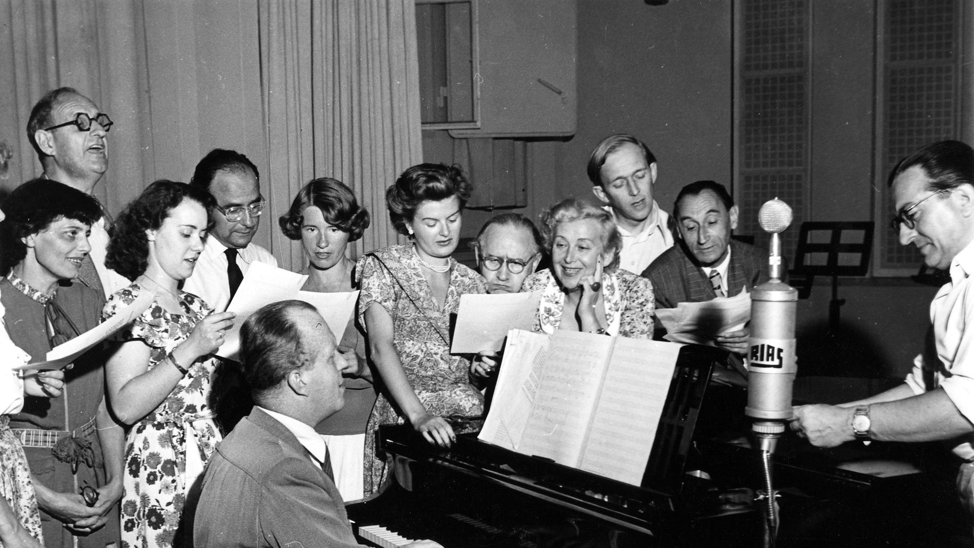 Eine schwarz-weiß Aufnahme aus dem Jahr 1950. Ein Mann spielt am Klavier und ist umgeben von einer Gruppe von Frauen und Männern, die währenddessen singt. 