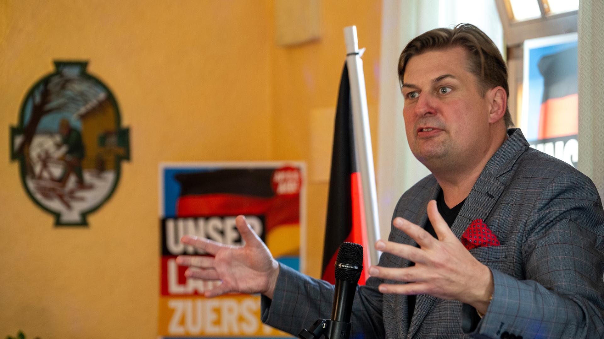 Krah spricht und gestikuliert mit beiden Händen in einem Raum. Im Hintergrund sind eine Deutschland-Fahne und ein AfD-Wahlplakat zu erkennen.