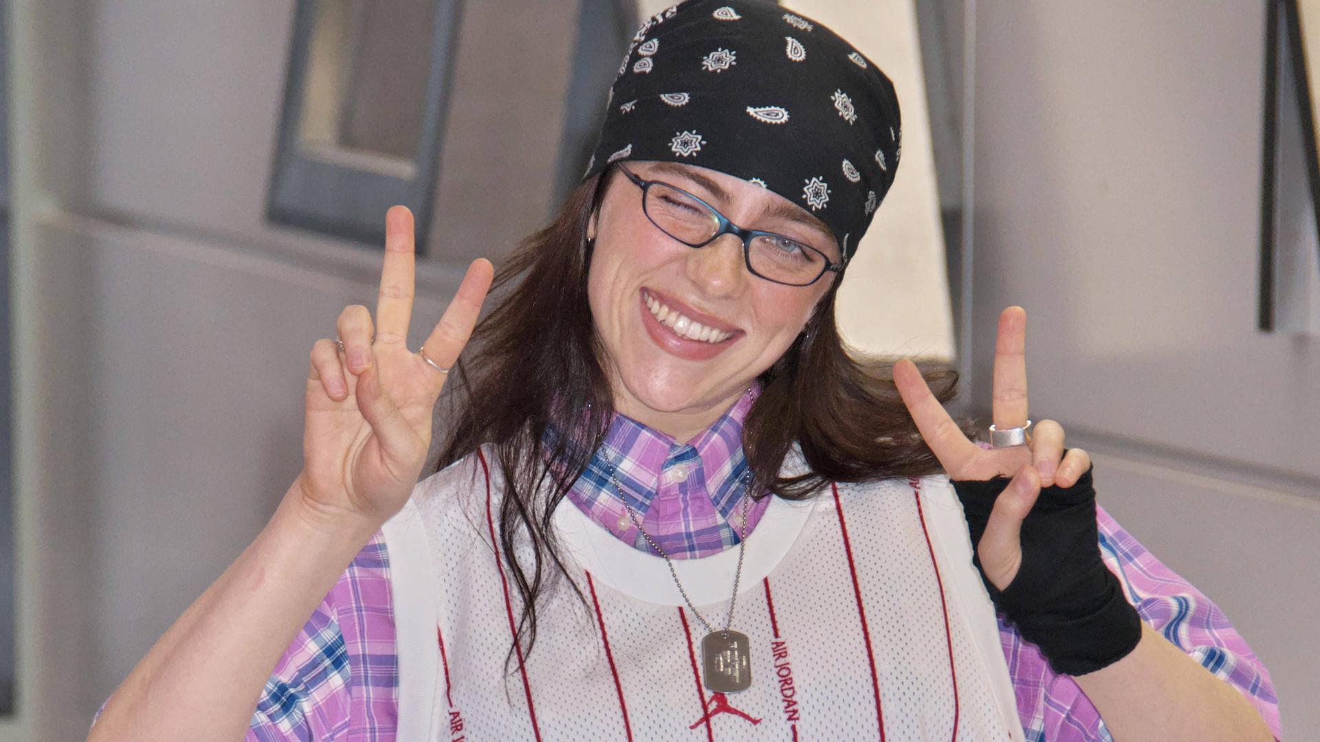 Musikerin Billie auf dem Narita International Airport in Japan. Sie trägt Brille und Kopftuch und zeigt mit beiden Händen das Victory-Zeichen. Musikerin Billie auf dem Narita International Airport in Japan. Sie trägt Brille und Kopftuch und zeigt mit beiden Händen das Victory-Zeichen.