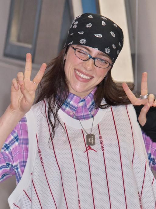Musikerin Billie Eilish auf dem Narita International Airport in Japan. Sie trägt Brille und Kopftuch und zeigt mit beiden Händen das Victory-Zeichen.
