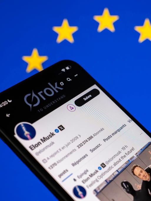 Eine Hand hält ein Mobiltelefon auf dessen Bildschirm der KI-Chatbot Grok der Plattform X zu sehen ist. 