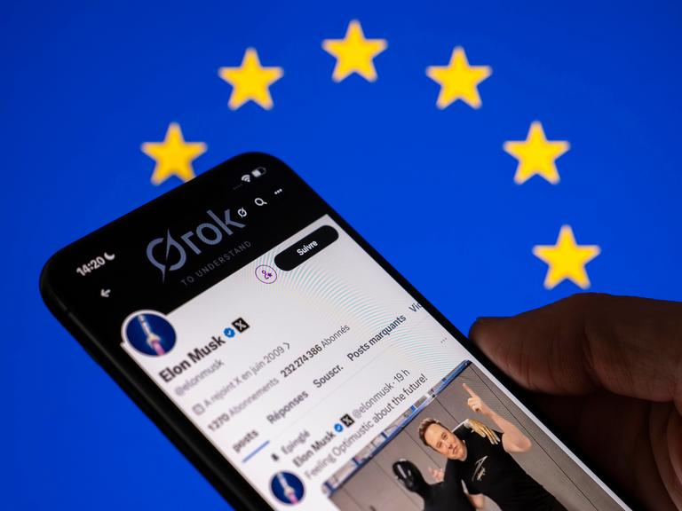 Eine Hand hält ein Mobiltelefon auf dessen Bildschirm der KI-Chatbot Grok der Plattform X zu sehen ist. 