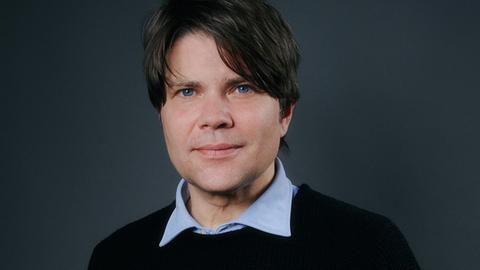 Ein Mann mit dunkelbraunem Haar und blauen Augen blickt in die Kamera. Er trägt einen schwarzen Pullover, über den der Kragen eines hellblauen Hemds ragt.