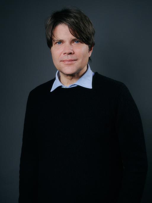 Ein Mann mit dunkelbraunem Haar und blauen Augen blickt in die Kamera. Er trägt einen schwarzen Pullover, über den der Kragen eines hellblauen Hemds ragt.