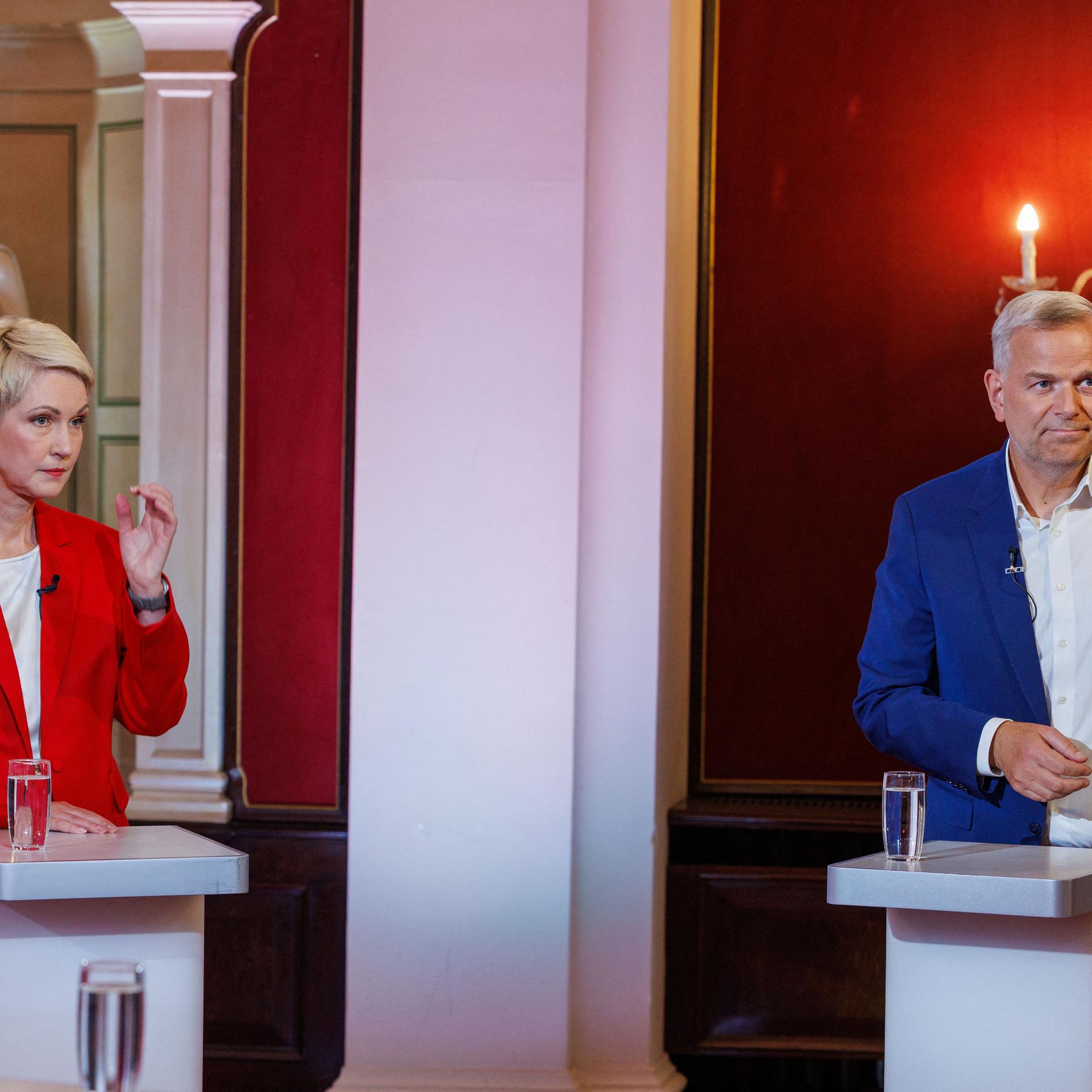 Manuela Schwesig und Leif-Erik Holm stehen bei einem TV-Duell deutlich voneinander getrennt an zwei Redepulten und reagieren auf die Fragen der Moderatorin. Parteifarbentypisch trägt sie ein rotes Kostüm und der einen blauen Anzug.