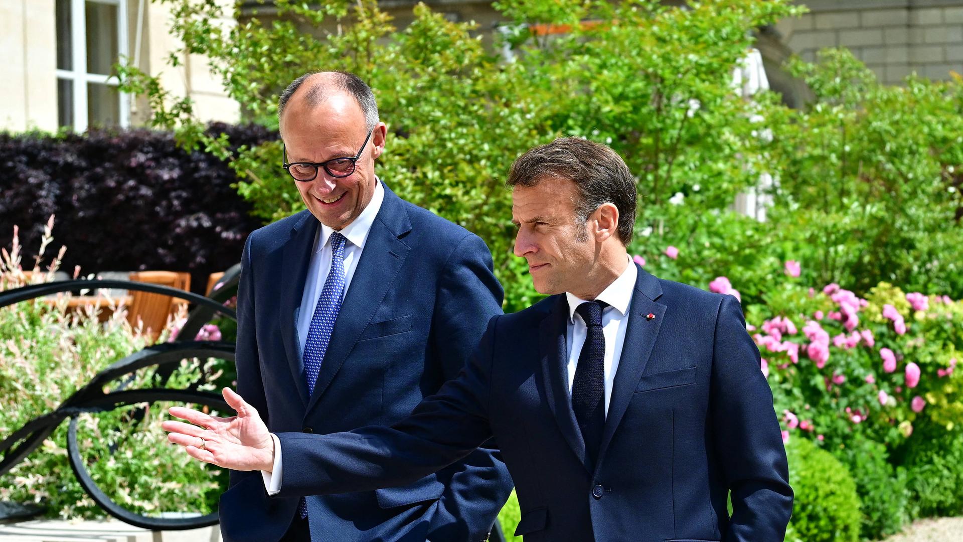 Friedrich Merz (links) und Emmanuel Macron (rechts) laufen gutgelaunt in dunklen Anzugen durch einen Park