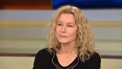 Irene Dische trägt ein schwarzes Oberteil, offene helle und gewellte Haare und sitzt in einem Fernsehstudio.