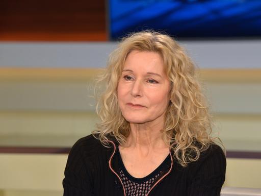 Irene Dische trägt ein schwarzes Oberteil, offene helle und gewellte Haare und sitzt in einem Fernsehstudio.