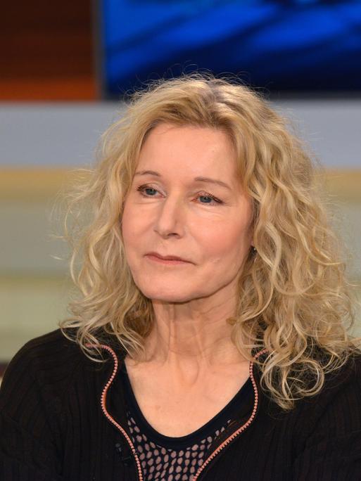 Irene Dische trägt ein schwarzes Oberteil, offene helle und gewellte Haare und sitzt in einem Fernsehstudio