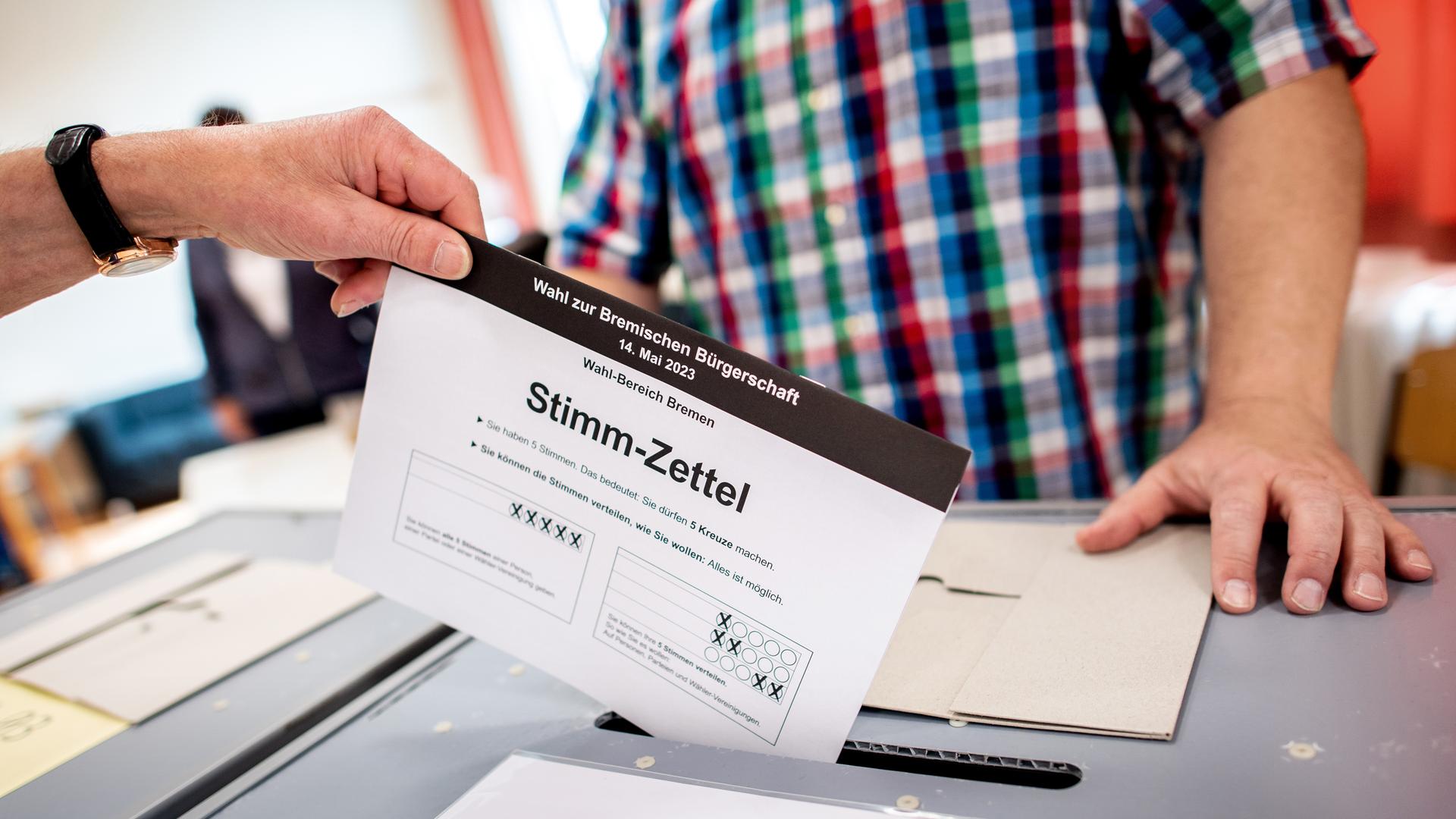 Eine Hand steckt einen Stimmzettel in eine Wahlurne. Im Hintergrund wacht ein Helfer über den korrekten Ablauf.