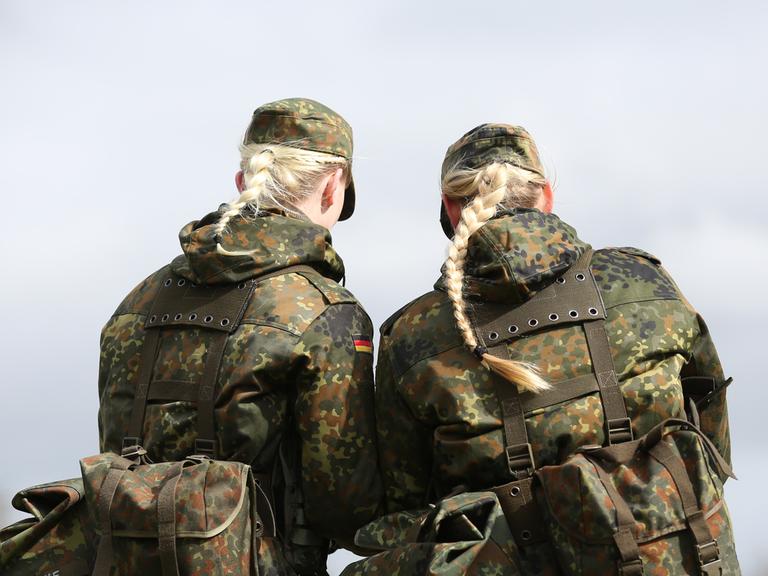 Das Foto zeigt Soldatinnen der Bundeswehr von der Hinterseite und in Tarnkleidung. Aus ihren Mützen hängt jeweils ein langer blonder Haarzopf heraus. 