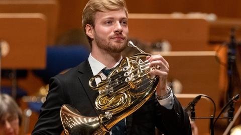 Ein junger Mann mit blonden Haaren steht mit seinem Horn auf der Bühne, kurz bevor er seinen Einsatz hat.