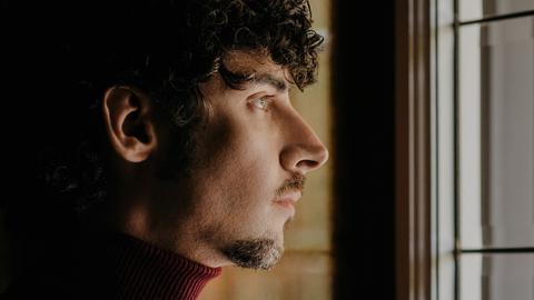 Ein junger Mann mit kurzen schwarzen Locken blickt aus einem Fenster. Wir sehen sein Gesicht im Profil. 