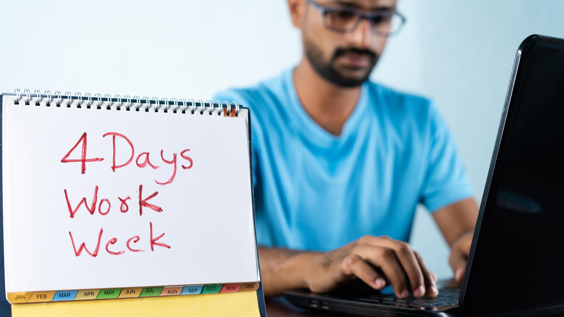 Ein Mann sitzt am Schreibtisch vor einem Laptop bei der Arbeit. Auf seinem Kalender hat er geschrieben "4 Days Work Week".