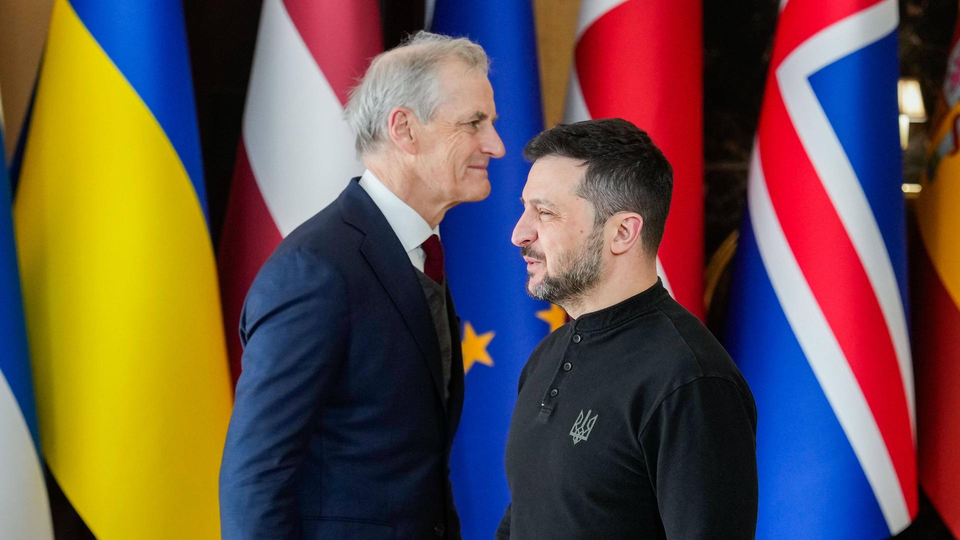 Der norwegische Premierminister Jonas Gahr Stoere und der ukrainische Präsident Volodymyr Selenskyj. Vor verschiedenen Flaggen gehen sie aneinander vorbei.