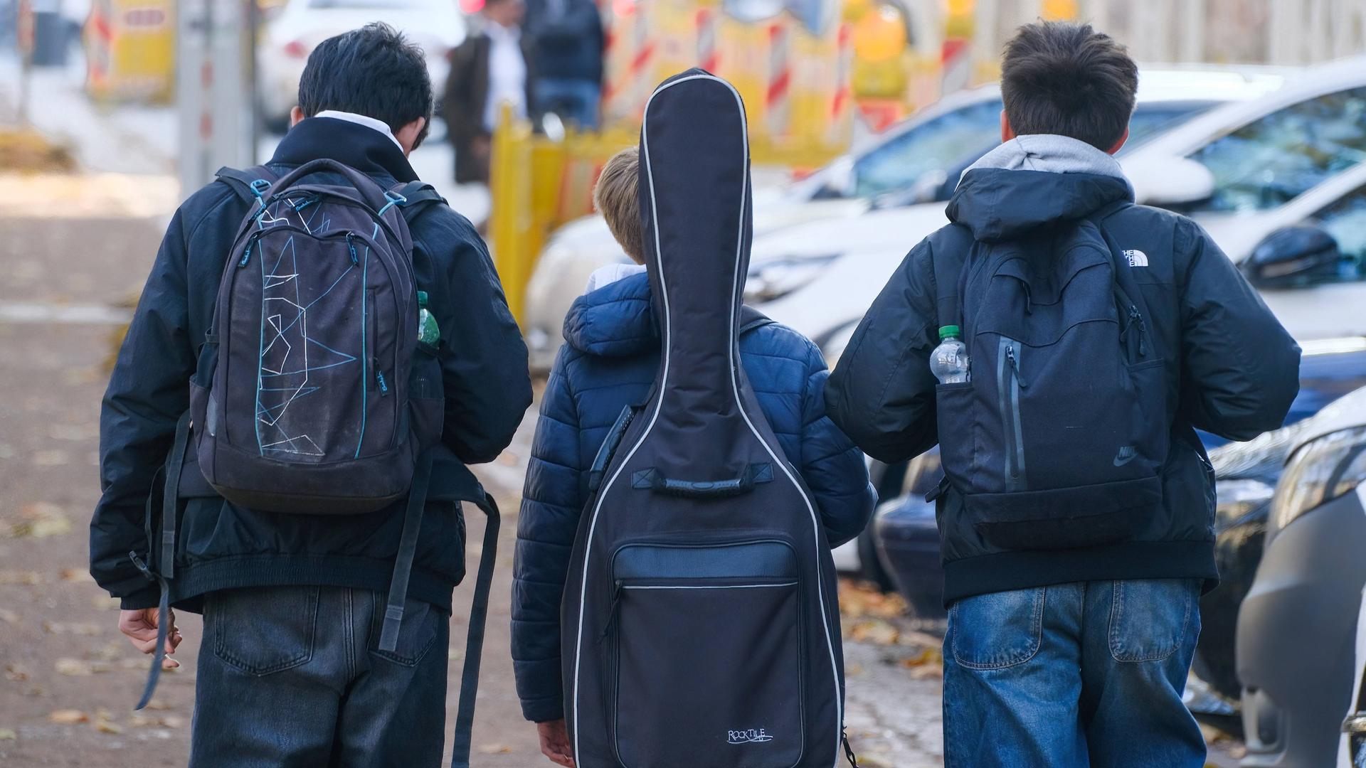 Das Bild zeigt drei männliche Jugendliche (aufgenommen von hinten). Die drei Jungen gehen nebeneinander auf einem Bürgersteig. Die beiden äußeren tragen Rucksäcke, die mittlere Person trägt eine Gitarrentasche auf dem Rücken.