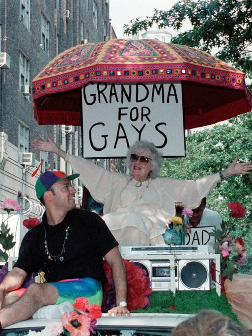 Beim CSD in New York 1989 sitzt eine ältere Dame unter einem Schild mit der Aufschrift "Grandma for Gays" und winkt den anderen Teilnehmern der Parade zu. Beim CSD in New York 1989 sitzt eine ältere Dame unter einem Schild mit der Aufschrift "Grandma for Gays" und winkt den anderen Teilnehmern der Parade zu.