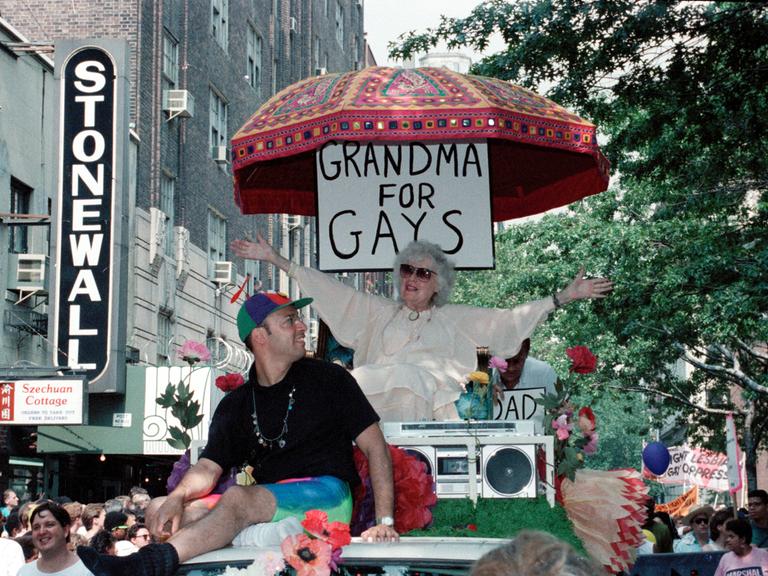 Beim CSD in New York 1989 sitzt eine ältere Dame unter einem Schild mit der Aufschrift "Grandma for Gays" und winkt den anderen Teilnehmern der Parade zu. Beim CSD in New York 1989 sitzt eine ältere Dame unter einem Schild mit der Aufschrift "Grandma for Gays" und winkt den anderen Teilnehmern der Parade zu.