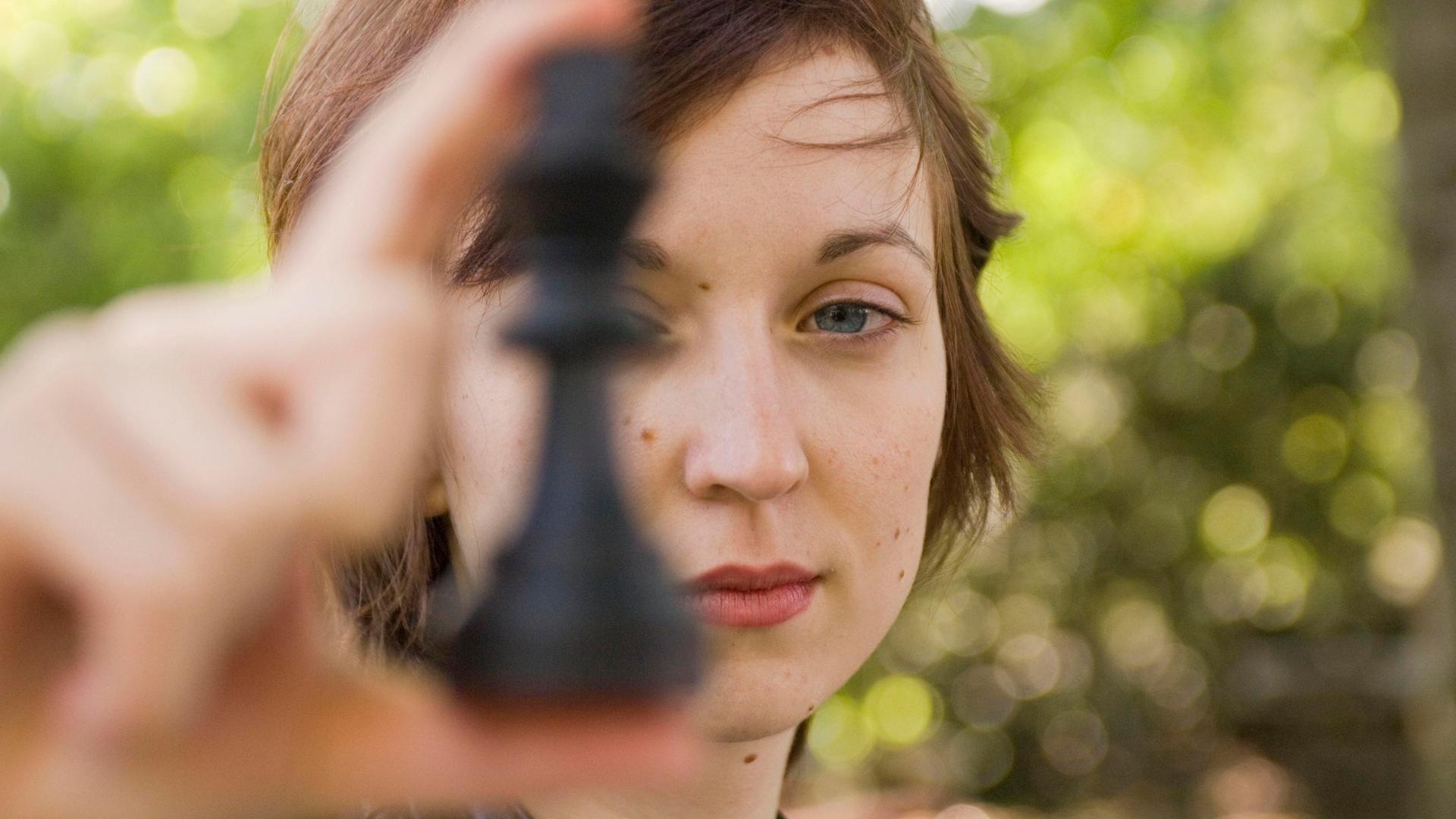 Porträt von Elisabeth Pähtz als Junioren-Weltmeisterin im Schach. Sie hält sich eine Schafigur vor das linke Auge und schaut dabei sehr ernst.