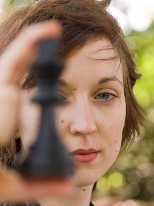 Porträt von Elisabeth Pähtz als Junioren-Weltmeisterin im Schach. Sie hält sich eine Schafigur vor das linke Auge und schaut dabei sehr ernst.