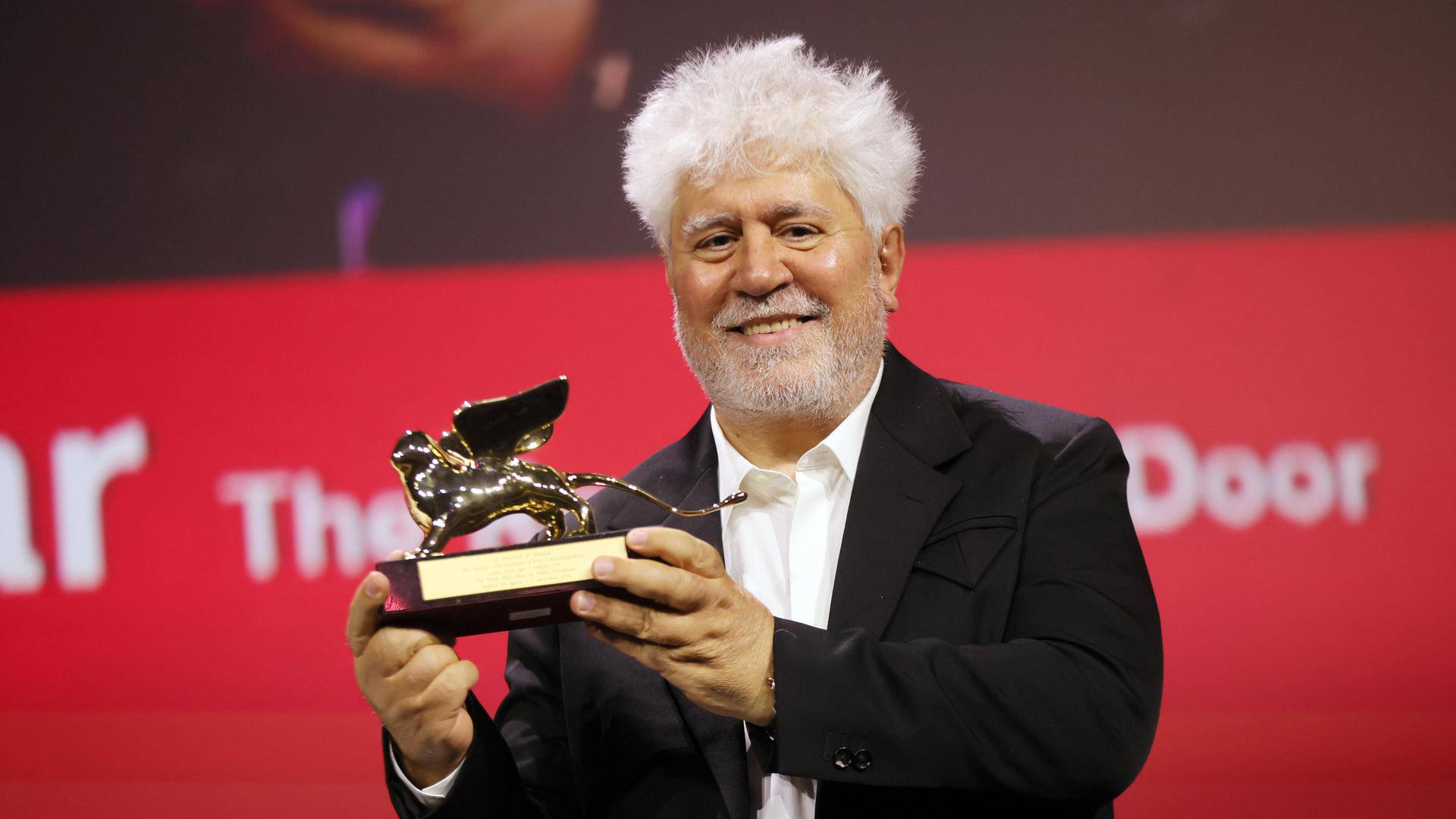 Der spanische Regisseur Pedro Almodóvar hält eine geflügelte Löwenstatue in den Händen und lächelt. 