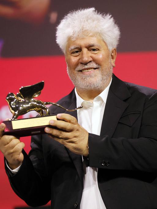 Der spanische Regisseur Pedro Almodóvar hält eine geflügelte Löwenstatue in den Händen und lächelt. 