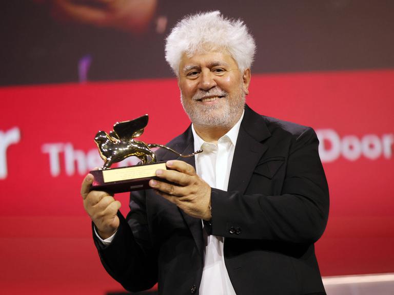 Der spanische Regisseur Pedro Almodóvar hält eine geflügelte Löwenstatue in den Händen und lächelt. 