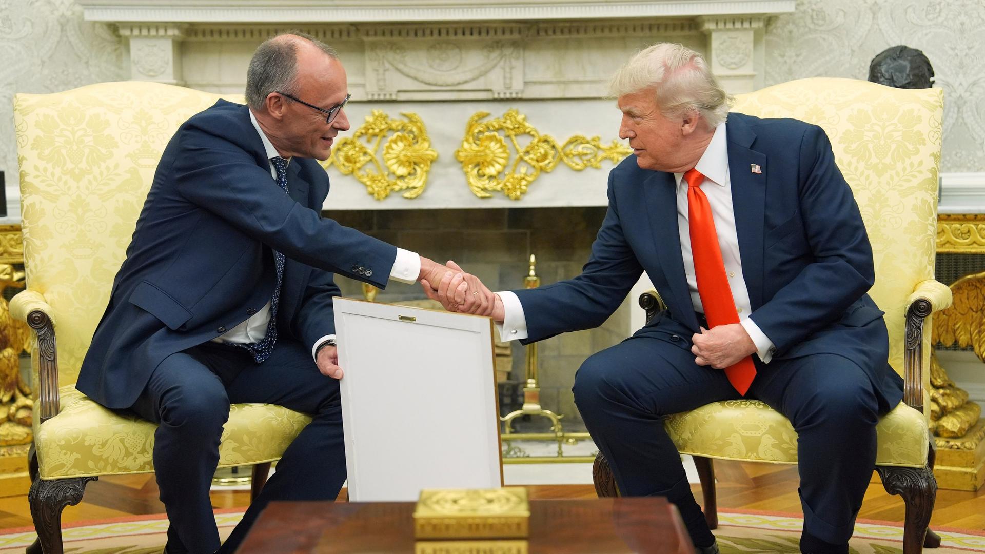 Bundeskanzler Friedrich Merz und US-Präsident Donald trump sitzen auf gelben Sesseln und schütteln sich die Hände.