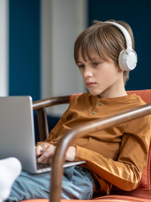 Ein Junge mit Kopfhörern sitzt mit Tablet auf dem Schoss in einem Sessel