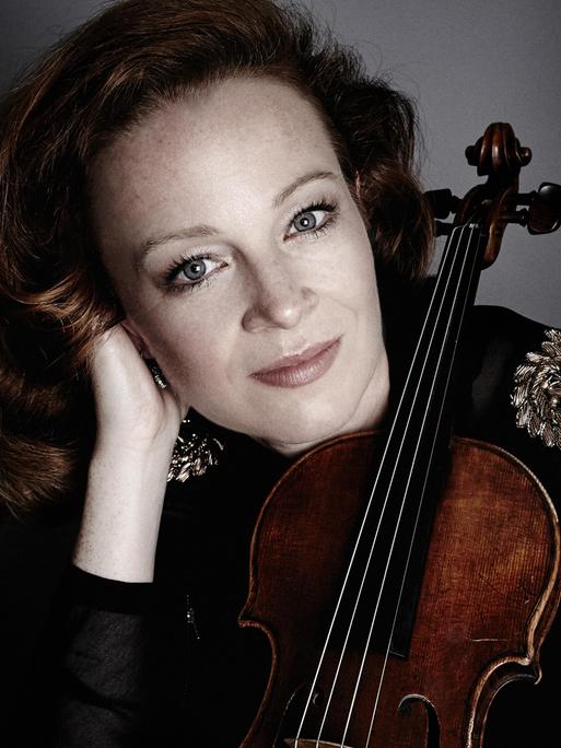 Eine Frau mit rotbraunen Haaren stützt ihren Kopf auf ihre rechte Hand. Gegen ihre linke Schulter lehnt eine Geige. Die Frau blickt freundlich.