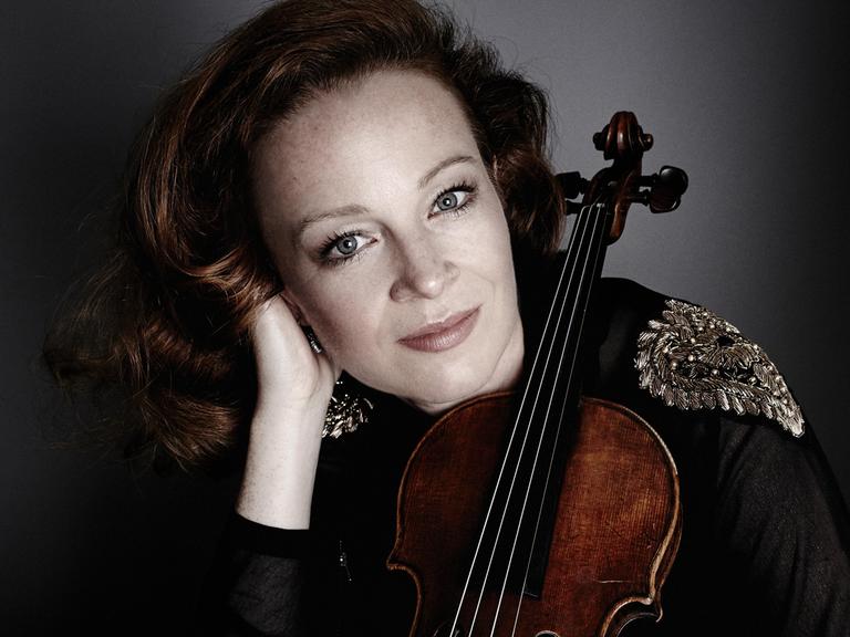 Eine Frau mit rotbraunen Haaren stützt ihren Kopf auf ihre rechte Hand. Gegen ihre linke Schulter lehnt eine Geige. Die Frau blickt freundlich.