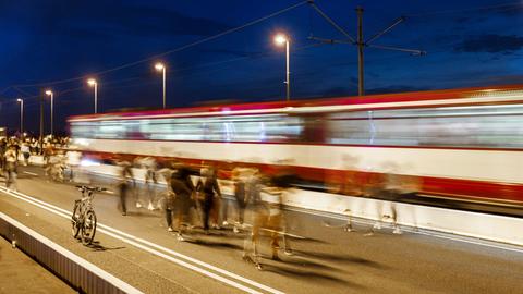 Das Bild zeigt Menschen, die abends über eine beleuchtete Brücke gehen. Bewusst eingesetzte Bewegungsunschärfe lässt die Menschen verschwimmen. Im Hintergrund fährt eine rot-weiße-Straßenbahn über die Brücke (ebenfalls verschwommen). 