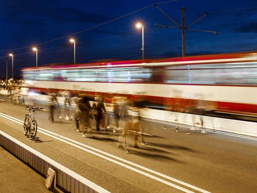 Das Bild zeigt Menschen, die abends über eine beleuchtete Brücke gehen. Bewusst eingesetzte Bewegungsunschärfe lässt die Menschen verschwimmen. Im Hintergrund fährt eine rot-weiße-Straßenbahn über die Brücke (ebenfalls verschwommen). 