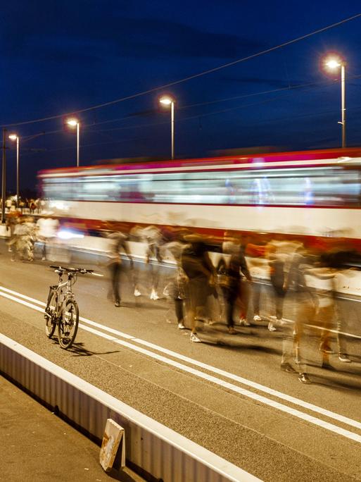 Das Bild zeigt Menschen, die abends über eine beleuchtete Brücke gehen. Bewusst eingesetzte Bewegungsunschärfe lässt die Menschen verschwimmen. Im Hintergrund fährt eine rot-weiße-Straßenbahn über die Brücke (ebenfalls verschwommen). 