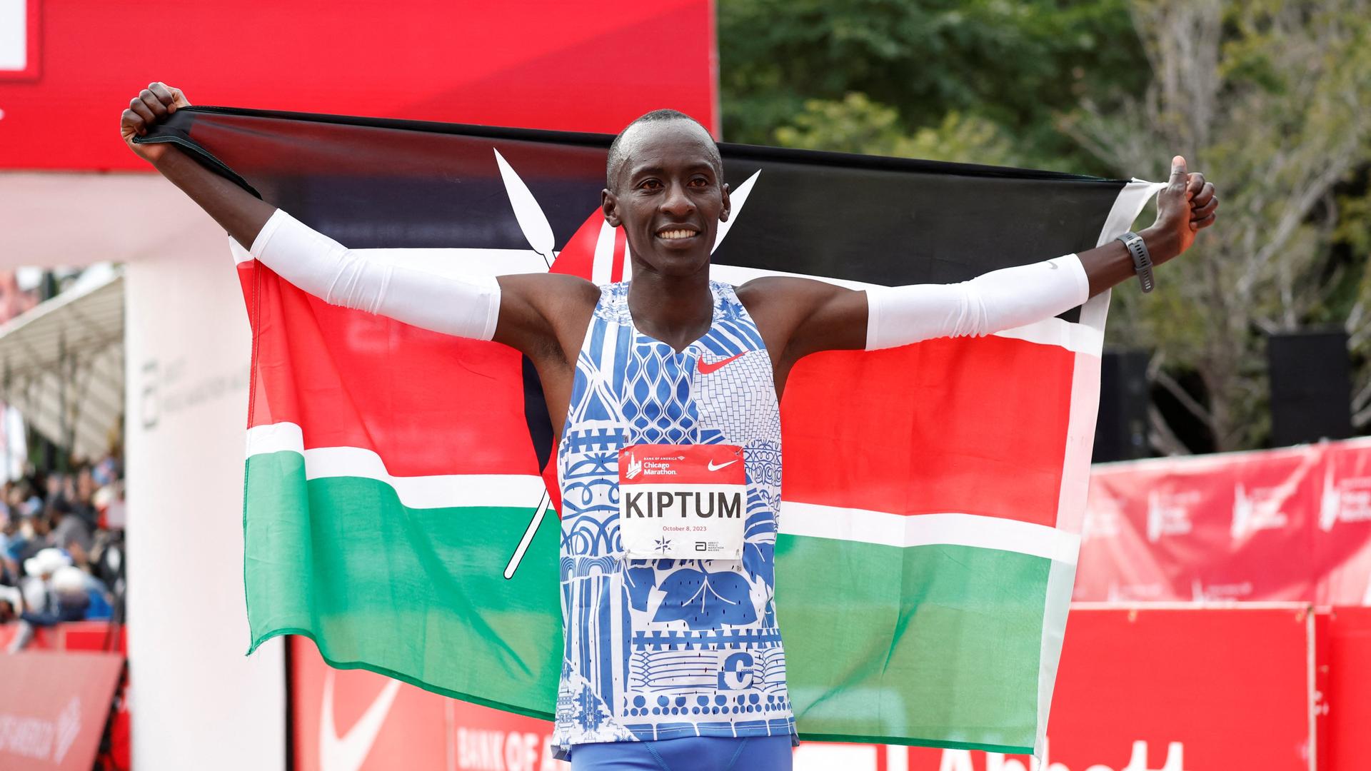 Kiptum hält eine kenianische Flagge hinter seinem Rücken in die Höhe. Er trägt ein hellblaues Trikot mit seinem Namen.