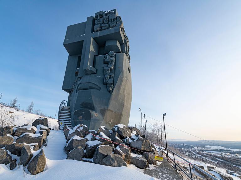 Eine großes, maskenähnliches Denkmal aus Stein mit christlichen Zeichen steht in einer Winterlandschaft.