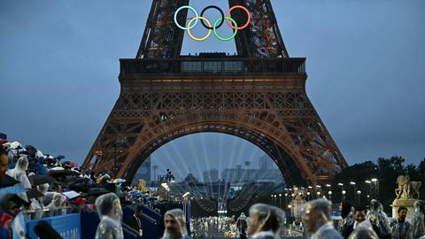 Sportlerinnen und Sportler in Regenkleidung vor dem Eiffelturm, der die Olympischen Ringe trägt. 